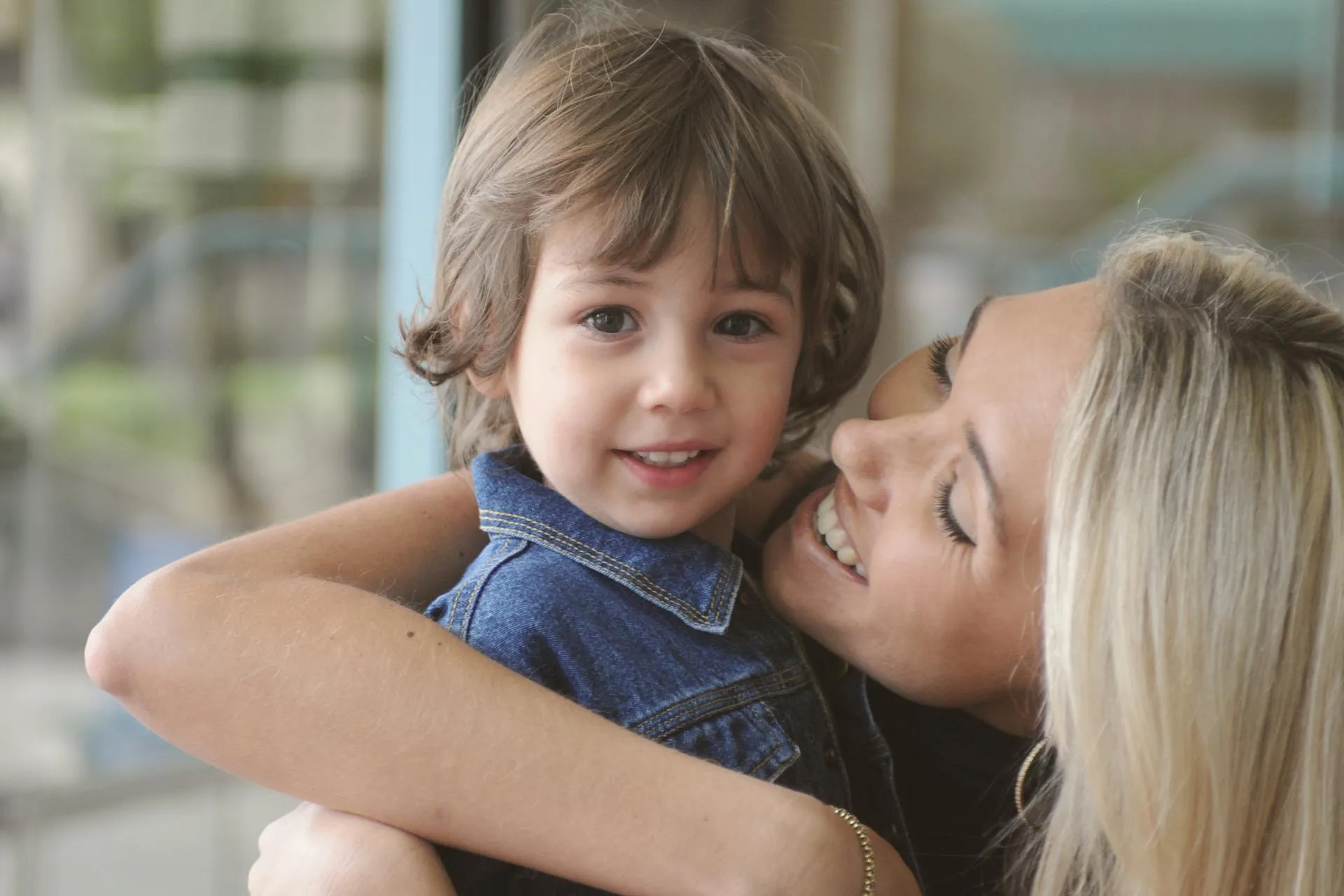 Smiling blonde woman hugging a young boy in a blue denim jacket, with the boy looking at the camera.